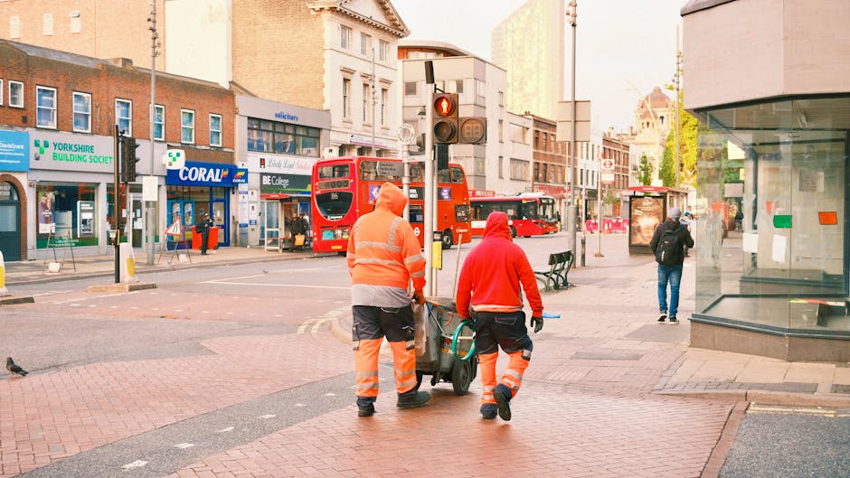 Two cleaning workers dressed in high-visibility orange and red uniforms are pushing a cleaning trolley on a city street in Tottenham High Road, Haringey. The background features a variety of urban elements including a red double-decker bus, storefronts for the Yorkshire Building Society, Coral, and BE College, and a traffic light showing red. The street is paved with bricks and surrounded by multi-story buildings with shop windows. The area appears clean and well-maintained, with some pedestrians walking along the sidewalk, and natural daylight illuminating the scene. [COMPANY_NAME] is providing surface cleaning and maintenance services in this urban environment, supporting the accessible and hygienic condition of the area.