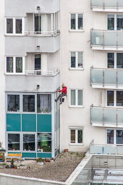 A window cleaner wearing a red jacket and safety harness is suspended on the exterior of a multi-storey residential building on Tottenham High Road, Haringey. The building features white walls, several glass balconies with metal railings, and large windows, some with partially visible interior furnishings. The windows are clean and reflective, with some having open curtains or blinds, indicating residential occupancy. The ground below shows a small landscaped area with gravel and potted plants. Sunlight illuminates the scene, highlighting the building's facade. This image relates to the services provided by Cleaners Haringey, including surface cleaning and deep cleaning of residential buildings, addressing access challenges for terrace cleaning, as shown in the context of the page titled 'Access problems for terrace cleaning on Tottenham High Road, Haringey.'