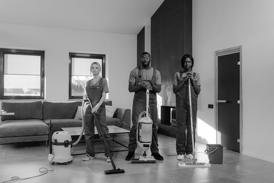 Three professional cleaners from Cleaners Haringey are performing surface cleaning and deep cleaning tasks in a modern living room. The room features a large, plush fabric sofa positioned against the wall beneath two windows, allowing natural light to illuminate the space. The floor is tiled with smooth, reflective ceramic tiles, which appear spotless and shiny, indicating thorough cleaning. One cleaner, standing in the middle, is holding a vacuum cleaner with a hose attachment, ready to service the carpeted areas or hard floors. To the right, another cleaner is using a mop and bucket, likely engaged in wet surface cleaning or sanitisation of the floor. The third cleaner on the left is holding a cloth or spray bottle, possibly mopping, wiping, or applying cleaning solutions to surfaces. Visible cleaning equipment, including vacuum cleaners and mops, suggests a comprehensive cleaning process. The room's layout is open, with a sectional sofa and a sleek, modern fireplace or entertainment unit against the wall. The natural light streaming in highlights the cleanliness and organisation. The image exemplifies professional domestic cleaning services offered by Cleaners Haringey to maintain hygienic, well-maintained living spaces, aligned with advice on avoiding hidden cleaning charges in Haringey quotes.