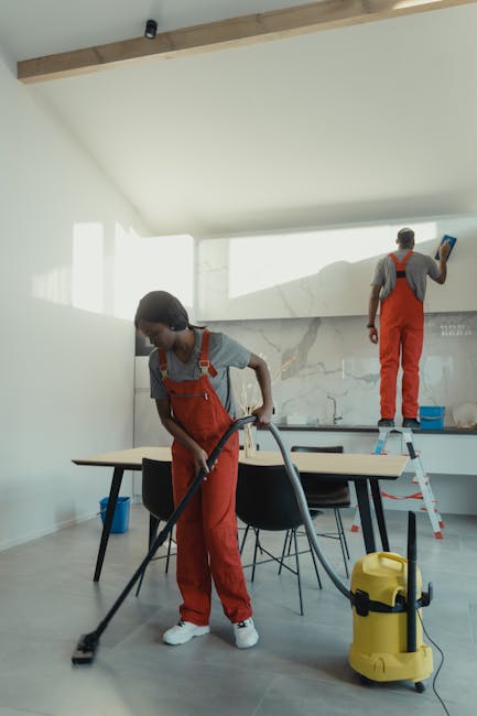 Two professional cleaners from Cleaners Haringey are engaged in surface cleaning within a modern, bright kitchen. The female cleaner in the foreground is using a vacuum or extractor to clean the tiled or hardwood floor, wearing red overalls and a grey t-shirt. In the background, a male cleaner is standing on a small ladder, wiping or polishing the tiled wall or backsplash with a cloth, also dressed in red overalls with a grey shirt underneath. The room features a sleek, contemporary design with white walls, a marble-patterned backsplash, and a large window providing ample natural light, highlighting the cleanliness and hygiene of the space. The minimalistic furniture includes a wooden table and black chairs, with cleaning tools and supplies visibly positioned around the area, emphasizing professional domestic cleaning processes. This image illustrates the comprehensive cleaning services offered by Cleaners Haringey, focusing on surface cleaning and maintenance in a residential setting, aligned with the page titled 'Avoid hidden cleaning charges in Haringey quotes, Haringey.'