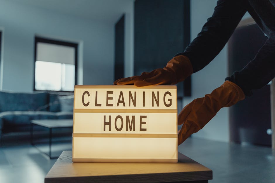 Three professional cleaners from Cleaners Haringey are performing surface cleaning and deep cleaning tasks in a modern living room. The room features a large, plush fabric sofa positioned against the wall beneath two windows, allowing natural light to illuminate the space. The floor is tiled with smooth, reflective ceramic tiles, which appear spotless and shiny, indicating thorough cleaning. One cleaner, standing in the middle, is holding a vacuum cleaner with a hose attachment, ready to service the carpeted areas or hard floors. To the right, another cleaner is using a mop and bucket, likely engaged in wet surface cleaning or sanitisation of the floor. The third cleaner on the left is holding a cloth or spray bottle, possibly mopping, wiping, or applying cleaning solutions to surfaces. Visible cleaning equipment, including vacuum cleaners and mops, suggests a comprehensive cleaning process. The room's layout is open, with a sectional sofa and a sleek, modern fireplace or entertainment unit against the wall. The natural light streaming in highlights the cleanliness and organisation. The image exemplifies professional domestic cleaning services offered by Cleaners Haringey to maintain hygienic, well-maintained living spaces, aligned with advice on avoiding hidden cleaning charges in Haringey quotes.