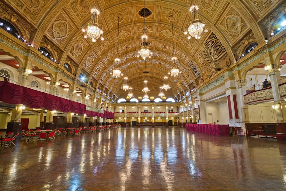 Interior view of a large, ornate hall with a high, decorated arched ceiling adorned with intricate moldings and multiple grand chandeliers hanging from above. The polished wooden floor reflects the ambient lighting, showcasing its well-maintained shine. Along the left side of the room, there are rows of red cushioned chairs and a deep red decorative curtain draped along the upper wall. The space is brightly lit with natural light streaming through large arched windows and supplemented by the chandeliers, highlighting the cleanliness and freshness of the surface areas. The overall appearance suggests that the hall has been thoroughly cleaned and maintained, with a focus on deep cleaning, surface sanitisation, and ensuring a pristine environment, as provided by Cleaners Haringey, specialists in domestic and commercial cleaning.