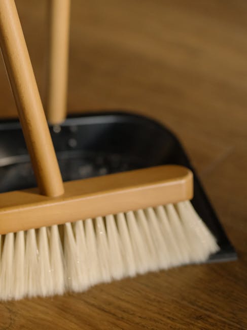 A close-up view of a broom and dustpan set on a wooden floor, with the broom's beige bristles and wooden handle prominently displayed. The dustpan is black with a wooden lip that matches the broom handle, positioned behind the broom. The scene highlights a clutter-free, clean floor surface prepared for sweeping, with soft, natural lighting emphasizing the smoothness of the wood. The background features a blurred wooden surface, suggesting a residential or commercial interior. Cleaners Haringey specializes in deep cleaning and surface sanitisation, ensuring thorough hygiene and maintained cleanliness within domestic or commercial spaces.