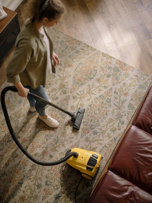 A woman is performing surface cleaning with a vacuum cleaner on a floral-patterned area rug in a living room. The rug, featuring intricate designs in muted colors, is positioned on a light wooden floor. She is dressed in casual clothing, including jeans, a beige jacket, and sneakers, and is holding the vacuum cleaner's hose with both hands. The room is well-lit, highlighting the clean and tidy appearance of the rug and the surrounding wooden floor area. In the background, there is a leather sofa partially visible. This scene demonstrates domestic cleaning services provided by Cleaners Haringey, focusing on deep cleaning and sanitisation of carpets and rugs.