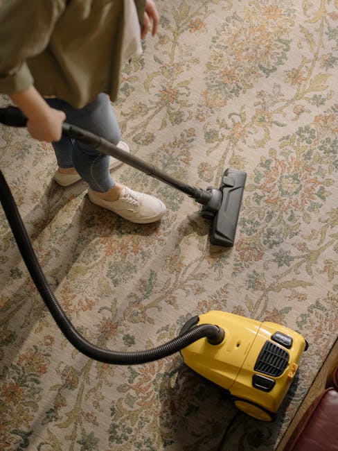 A woman is performing surface cleaning with a vacuum cleaner on a floral-patterned area rug in a living room. The rug, featuring intricate designs in muted colors, is positioned on a light wooden floor. She is dressed in casual clothing, including jeans, a beige jacket, and sneakers, and is holding the vacuum cleaner's hose with both hands. The room is well-lit, highlighting the clean and tidy appearance of the rug and the surrounding wooden floor area. In the background, there is a leather sofa partially visible. This scene demonstrates domestic cleaning services provided by Cleaners Haringey, focusing on deep cleaning and sanitisation of carpets and rugs.