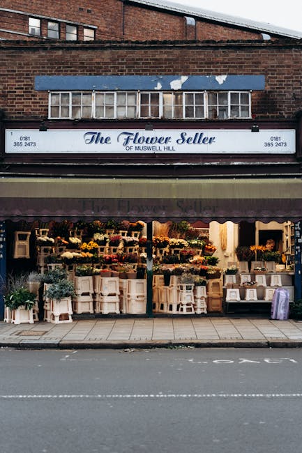The exterior of 'The Flower Seller' shop in Muswell Hill, featuring a glass-fronted display window with flower arrangements and potted plants arranged on wooden shelves and tables. The storefront has a maroon awning and a blue sign with white lettering. The building's upper portion is brick with a row of windows, and the sidewalk in front is clean and unobstructed. Natural daylight illuminates the scene, highlighting the neat presentation of floral displays, indicative of a well-maintained business that values cleanliness and presentation. The image reflects a typical commercial space that may require surface cleaning and sanitisation as part of maintenance from professionals like Cleaners Haringey, aligning with their expertise in domestic and commercial cleaning services.